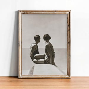 Op de afbeelding: Zwart-wit ingelijste foto van een stel in vintage badkleding, zittend en tegenover elkaar. De foto staat in een goudkleurige lijst, tegen een witte achtergrond en een houten oppervlak.