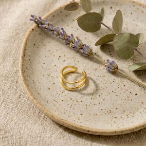 May include: A gold double-band ring on a speckled beige plate, with lavender and eucalyptus. The ring has a polished finish. The plate is on a neutral linen surface. The ring is a piece of jewellery.
