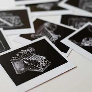 Peut inclure: Plusieurs tirages de style polaroid en noir et blanc représentant des illustrations détaillées d'appareils photo anciens. Les tirages mettent en valeur différents modèles d'appareils photo, en insistant sur leurs conceptions complexes et leurs éléments mécaniques. Les images sont disposées sur une surface claire.