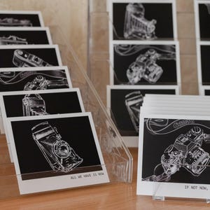 Peut inclure: Des tirages d'art en noir et blanc de vieux appareils photo sont exposés dans un présentoir en acrylique transparent. Les tirages présentent des dessins au trait détaillés d'appareils photo sur fond noir. Le texte indique "ALL WE HAVE IS NOW" et "IF NOT NOW, THEN WHEN?"