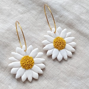 Puede incluir: Un par de pendientes de margarita. Cada pendiente presenta una flor de margarita blanca con un centro amarillo, suspendida de un aro dorado. Los pendientes están colocados sobre un fondo de tela neutra.