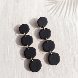May include: A pair of drop earrings featuring four matte black, rounded hexagonal shapes connected by small gold-toned metal jump rings. The earrings are arranged in a vertical column, creating a linear drop design with a textured surface finish.