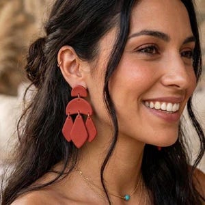 Può includere: Un paio di orecchini pendenti color terracotta caratterizzati da una parte superiore a semicerchio collegata a tre pendenti allungati a forma di diamante. Gli orecchini hanno una finitura opaca e sono appesi a un piccolo gancio.