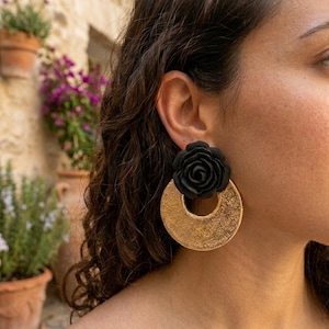 Puede incluir: Un pendiente llamativo que presenta una rosa esculpida en negro mate unida a un gran colgante circular de tono dorado con un acabado de metal martillado y texturizado. El pendiente está diseñado con un cierre de poste y cuelga verticalmente.