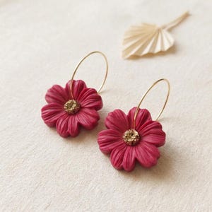 Puede incluir: Un par de pendientes de aro con colgantes en forma de flor de color magenta intenso. Cada flor tiene pétalos texturizados y un centro circular con un acabado dorado granulado y brillante. Aros finos de metal dorado en la parte superior.