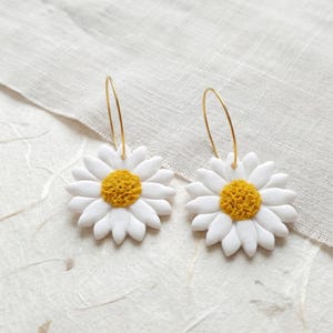 Puede incluir: Un par de pendientes de margarita. Cada pendiente presenta una flor blanca con un centro amarillo y un aro dorado. Los pendientes están colocados sobre un fondo neutro.