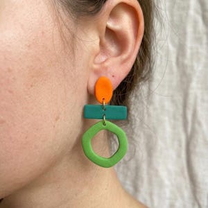Puede incluir: Un pendiente colgante que presenta tres formas geométricas apiladas: un óvalo naranja sólido en la parte superior, una barra rectangular de color verde azulado en el medio y un anillo hexagonal abierto de color verde lima en la parte inferior, unidos por anillas doradas.