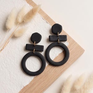 Puede incluir: Un par de pendientes negro mate con un diseño geométrico. Cada pendiente consta de una pieza superior hexagonal pequeña conectada a una barra rectangular horizontal, unida a un gran aro circular en la parte inferior con anillas doradas.