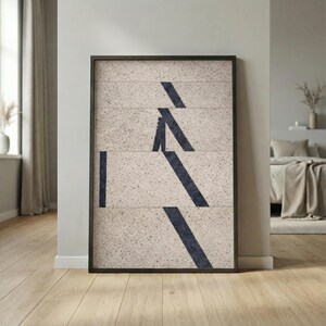 May include: Framed abstract art print featuring a beige speckled background with dark blue geometric shapes. The artwork is encased in a black frame and leans against a light gray wall, showcasing a modern aesthetic.