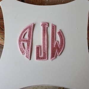 May include: White wooden decorative piece with a pink glitter monogram of the letters AJW. The monogram is in a circular design. The wooden piece has a curved shape.