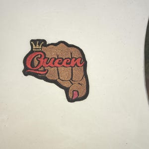 May include: A brown fist with a gold crown and the word "Queen" in red lettering. The fist is raised in the air.