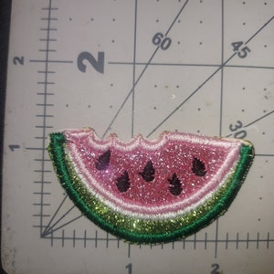 May include: A pink watermelon slice with green rind and black seeds. The watermelon is embroidered with glitter and has a white outline.