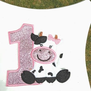 May include: A white applique featuring a smiling cow with black spots, a pink bow, and a black and pink hat. A large pink glittery number 1 is next to the cow. The design is on a white surface.