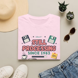 May include: A light pink t-shirt featuring a graphic with two floppy disks, a coffee cup, a loading bar, and the text "STILL PROCESSING SINCE 1983" in pink and green. A yellow sticky note with the text "Maybe Tomorrow" is also printed on the shirt.