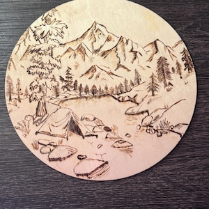May include: Circular wooden artwork with a camping theme. The pyrography art shows a mountain range, trees, a tent, and a river with rocks. The artwork is burned into the wood, creating a detailed, rustic aesthetic.
