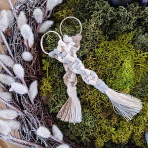 May include: Two macrame keyrings with silver rings. The keyrings are braided with grey, beige, and white cords, and have matching tassels. The background features green moss and a wreath with white fluffy accents.