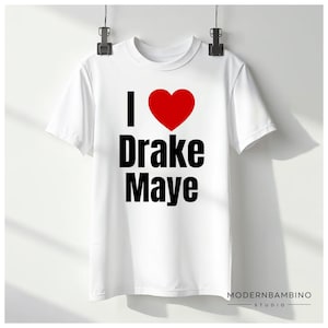 May include: White t-shirt with the text "I ❤️ Drake Maye" in black and a red heart. The shirt is hanging from a black clip. The Modern Bambino Studio logo is at the bottom right.