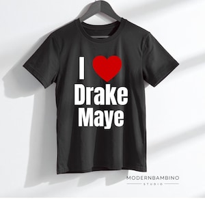 May include: Black t-shirt with the text "I ❤️ Drake Maye" in white and a red heart graphic. The shirt is hanging on a black hanger against a white background. The ModernBambino Studio logo is at the bottom.