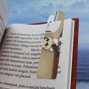 Puede incluir: Un marcapáginas con una ilustración de un hámster sosteniendo un arma, insertado en un libro. El marcapáginas tiene un fondo beige. El libro está abierto, con texto visible en las páginas. El fondo es un cielo azul.