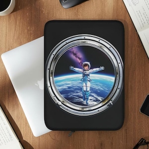 Puede incluir: Funda negra para portátil con un diseño de ventana circular que muestra a un astronauta flotando en el espacio. El astronauta lleva un traje espacial blanco y azul con los brazos extendidos. El fondo muestra la Tierra y una nebulosa púrpura.