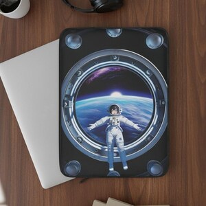 Puede incluir: Funda negra para portátil con un diseño de ventana circular que muestra a un astronauta con un traje espacial sobre un fondo de la Tierra y el espacio. La funda está sobre una superficie de madera con un portátil, una taza de café y auriculares.