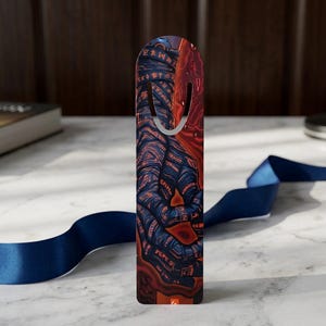 Puede incluir: Un marcapáginas vertical con un diseño gráfico en azul oscuro y rojo, con patrones intrincados y arremolinados y símbolos de color naranja brillante. El marcapáginas tiene un borde superior redondeado con una ranura y una cinta azul marino.