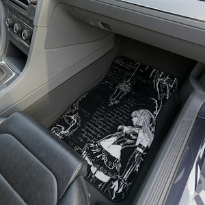 Könnte beinhalten: Schwarze Auto-Fußmatte mit einer weißen Anime-Illustration einer Frau mit langen Haaren, einem Gothic-Outfit und einem Schwert. Die Matte hat einen dekorativen Rand und schriftartähnlichen Text in Weiß.