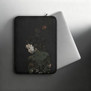 Puede incluir: Una funda para portátil negra con una ilustración detallada de una figura con cabello oscuro adornado con intrincados patrones florales y follaje. El diseño incorpora acentos rojos y blancos sobre un fondo oscuro, con cierre de cremallera.