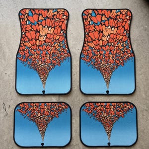 May include: A set of four car floor mats featuring a graphic print of a person standing at the bottom, with a large, dense swarm of orange and red butterflies rising upward against a solid light blue background, all bordered in black.