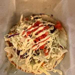 May include: A close-up of a taco on a white paper. The taco has a flatbread base, topped with shredded cabbage, red tomatoes, and a white sauce. The taco is in a metal container.