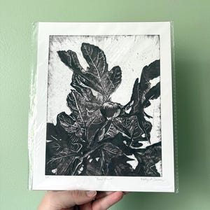 May include: Black and white linocut print of a fig tree branch with large leaves and a fig. The print is titled "Bear Fruit" and is signed by the artist. The print is in a clear plastic sleeve.