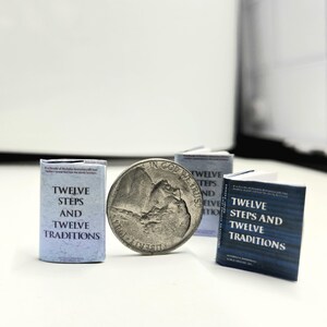 May include: A collection of miniature books and a coin. The books are titled "Twelve Steps and Twelve Traditions" in blue and white. The coin is a silver nickel with the words "In God We Trust".