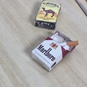 May include: Miniature cigarette packs of Camel Filters and Marlboro. The Camel pack has a yellow background with a camel and palm trees. The Marlboro pack is white with a red top and the brand name in bold, black letters.