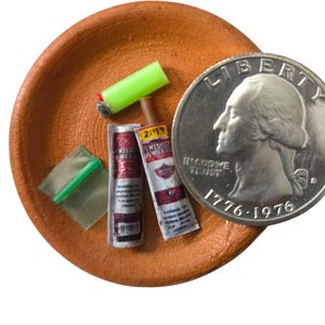 May include: A miniature terracotta-colored dish containing a tiny neon green lighter, two small Swisher Sweets cigarillo packages, and a small clear plastic bag with a green seal, positioned next to a United States quarter coin for scale.