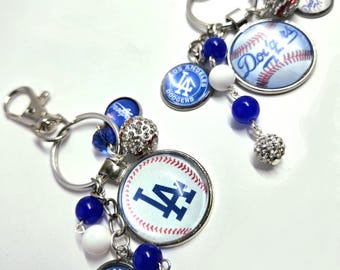 May include: Two Los Angeles Dodgers keychains featuring silver-tone metal rings and lobster clasps. Each keychain includes a round charm with the Dodgers logo, a baseball-themed charm, blue and white beads, and a sparkling silver rhinestone bead.