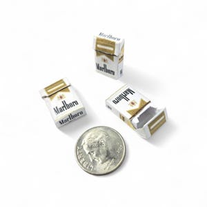 Puede incluir: Miniaturas de paquetes de cigarrillos Marlboro blancos y dorados con el nombre de la marca impreso. Un paquete está abierto, revelando el interior. Una moneda está al lado de los paquetes para la comparación de tamaño.