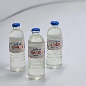 May include: Three miniature Evian water bottles with blue caps and labels. The labels feature the brand name and a mountain graphic. The bottles are clear, filled with liquid, and appear to be made of plastic.