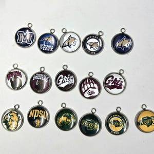 Peut inclure: Une collection de breloques rondes en métal avec divers logos et mascottes universitaires. Les breloques ont une bordure argentée et une boucle en haut pour l'attache. Les logos incluent "M", "Montana State", "Grizzlies" et "NDSU".