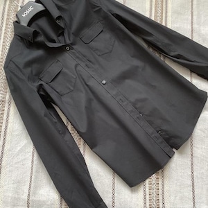 Pode incluir: Uma camisa preta com botões e dois bolsos no peito. A camisa de manga comprida tem punhos abotoados. A camisa é feita de tecido liso e é exibida em um cabide.
