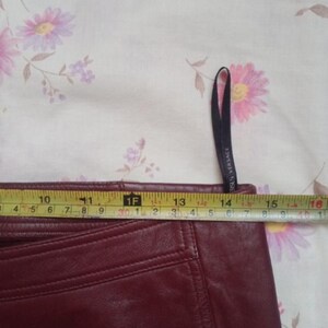 May include: A close-up of a burgundy leather skirt with a measuring tape showing a width of approximately 15 inches. The skirt has visible stitching and a tag that says "VERSACE". The background is a floral pattern.