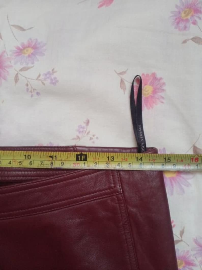 May include: A close-up of a burgundy leather skirt with a measuring tape showing a width of approximately 15 inches. The skirt has visible stitching and a tag that says "VERSACE". The background is a floral pattern.
