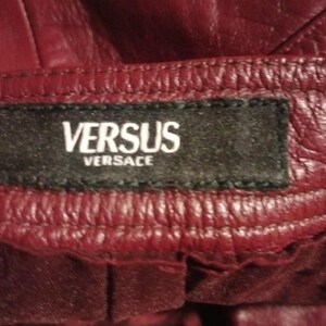 May include: Close-up of a burgundy leather garment with the brand label "VERSUS Versace" stitched on a black tag. The leather has a textured appearance with visible stitching.