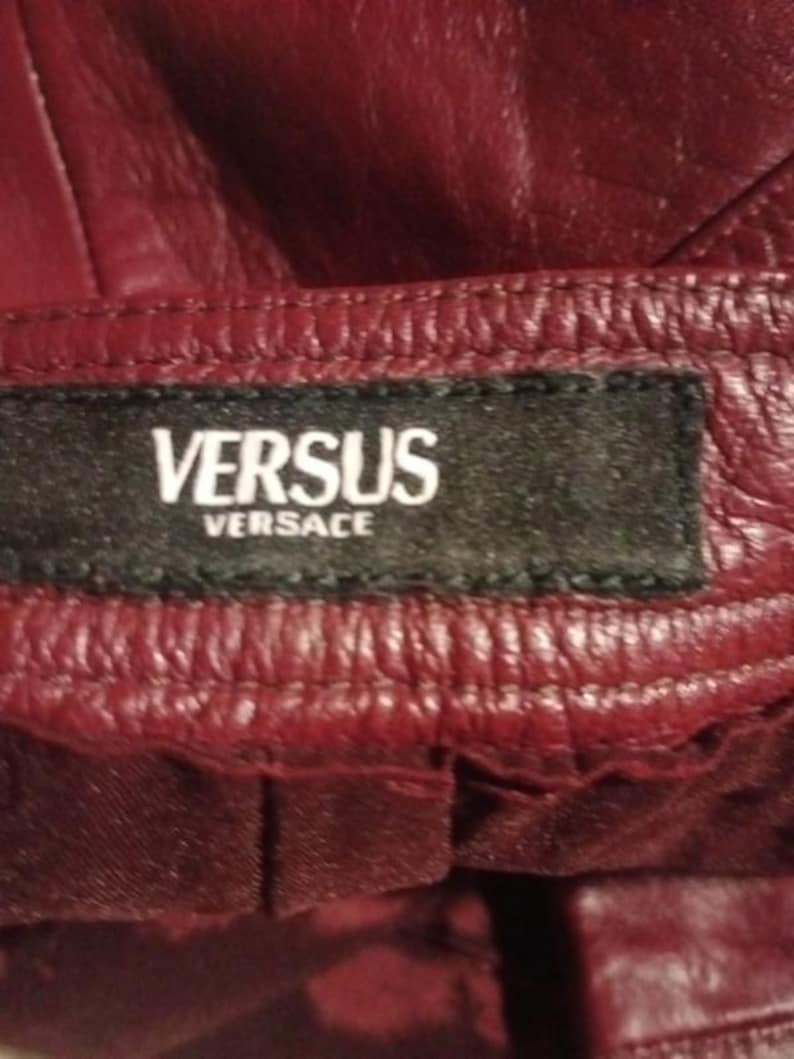 May include: Close-up of a burgundy leather garment with the brand label "VERSUS Versace" stitched on a black tag. The leather has a textured appearance with visible stitching.