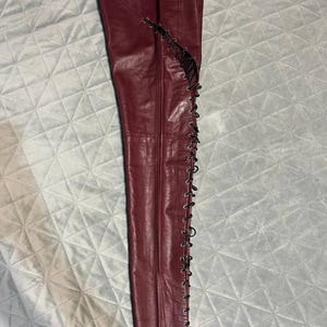 May include: Burgundy leather pants with a lace-up design along the outer seams. The pants are made of a smooth, shiny leather material and have a classic five-pocket design. The lace-up detail adds a unique and edgy touch to the pants.