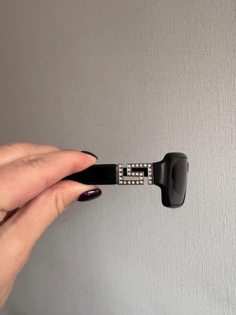 May include: Black sunglasses with a rectangular frame and dark lenses. The temples feature a silver-colored, geometric design encrusted with small, clear rhinestones. The sunglasses are held against a neutral gray background.