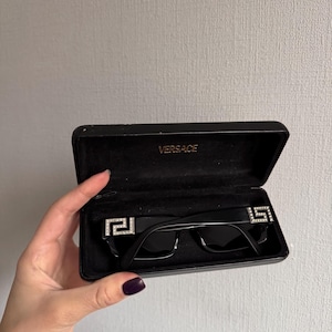 May include: Black sunglasses with a rectangular frame, displayed in an open black case. The sunglasses feature a silver emblem on each temple. The case is labeled "VERSACE" in gold lettering.