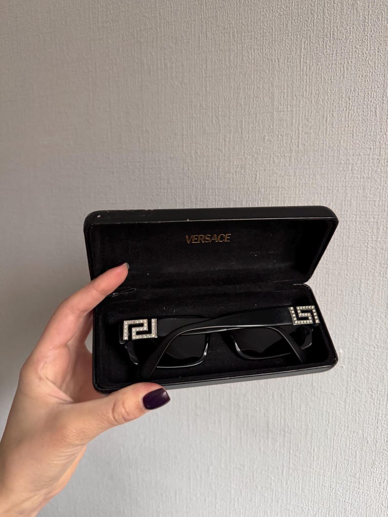 May include: Black sunglasses with a rectangular frame, displayed in an open black case. The sunglasses feature a silver emblem on each temple. The case is labeled "VERSACE" in gold lettering.