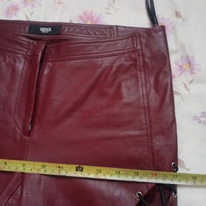 May include: Burgundy leather pants with a straight leg design. The pants feature a side lace-up detail and a small VERSUS label. A measuring tape shows a length of approximately 18 inches.