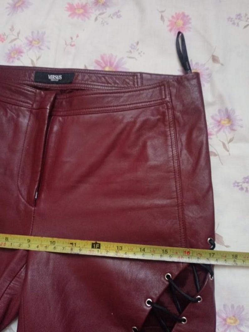 May include: Burgundy leather pants with a straight leg design. The pants feature a side lace-up detail and a small VERSUS label. A measuring tape shows a length of approximately 18 inches.