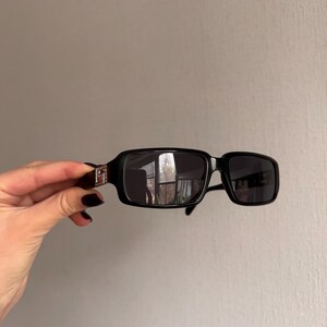 May include: Black rectangular sunglasses with dark lenses. The sunglasses have a thick frame and a silver accent on the temples. The lenses reflect the surrounding environment. The sunglasses are held by a hand.